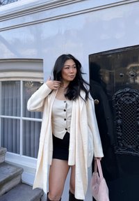 Cardigan crema sopra una camicia beige con bottoni, abbinato a una gonna nera e stivali alti marroni. Una borsa rosa pallido viene tenuta in una mano.