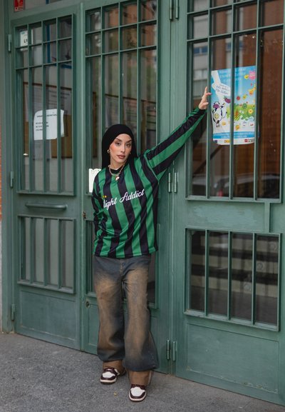 Camisa de manga larga a rayas verdes y negras con el texto "Light Addict", combinada con jeans de pierna ancha descoloridos y zapatillas blancas. De pie junto a puertas verdes.