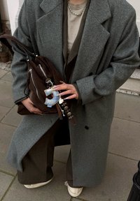 Grijze wollen overjas, bruine suède tas met ritsen, lichtgrijze top en bruine broek; met een blauw geruit letterbedeltje aan de tas.