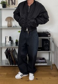 Zwarte bomberjack met een rits aan de voorkant, elastische mouwen en twee zijzakken. Gecombineerd met losse donkere jeans en witte schoenen met zwarte strepen.