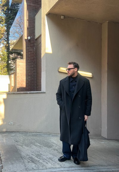 Abrigo negro de doble faz, cuello alto negro, pantalones azules y zapatos negros brillantes. Sosteniendo una bolsa oscura, de pie contra una pared texturizada.