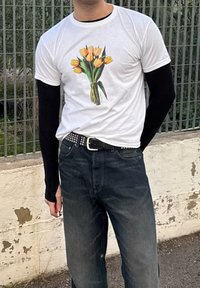 Λευκό μπλουζάκι με γραφικό μιας ανθοδέσμης από κίτρινες τουλίπες, φορεμένο πάνω από μακριά μαύρα μανίκια. Συνδυασμένο με σκούρα τζιν παντελόνια και μια ζώνη με καρφιά.