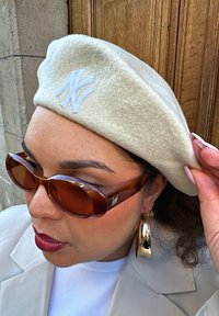 Beret bej din fetru cu logo brodat albastru, asortat cu ochelari de soare mari de culoare chihlimbar și cercei aurii în formă de cerc, pe un fundal de lemn.