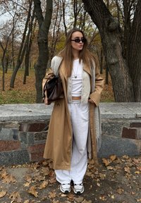 Tânără purtând o haină bej de tip trench, pantaloni sport albi, adidași și ochelari de soare, stă lângă un zid de piatră într-un parc cu frunze de toamnă.