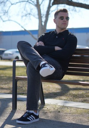 Joven con gafas de sol y ropa negra sentado con las piernas cruzadas en un banco de madera al aire libre, con los brazos cruzados, mirando de lado.