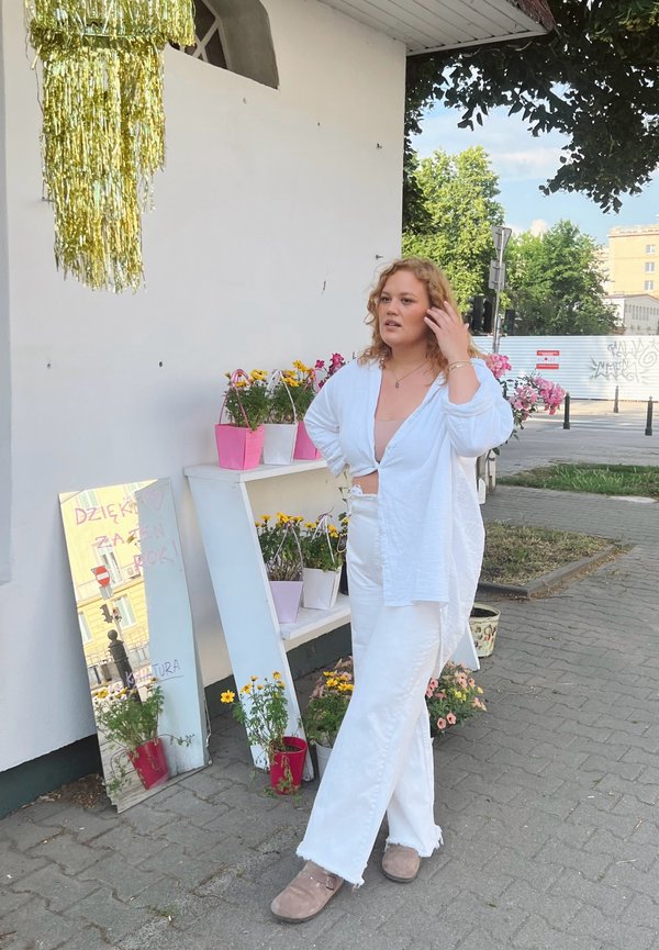Camisa de lino blanca, pantalones de denim blancos, calzado beige. Flores alrededor en macetas rosas y blancas, espejo reflectante mostrando texto.