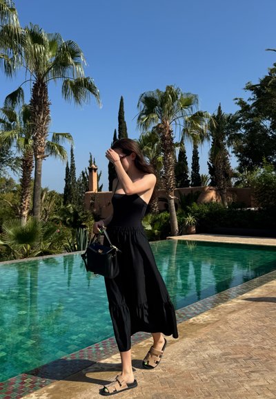 Vestido negro sin mangas con un bodice ajustado y una falda acampanada, combinado con sandalias color tierra. Los accesorios incluyen un bolso negro. Cielo azul despejado y palmeras.
