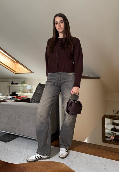 Mujer joven de pie en interiores, vistiendo un cárdigan morado oscuro, jeans de pierna ancha grises, zapatillas blancas y sosteniendo un pequeño bolso morado oscuro.