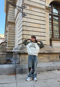 Vīrietis stāv uz ietves, labojot krēmbaltu un olīvu krāsas džemperi ar uzrakstu “Local Heroes”, valkājot platas, saplēstas džinsu bikses un baltas sporta kurpes.