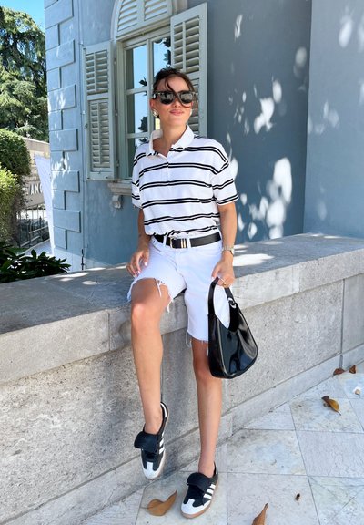 Camisa polo blanca con rayas negras, pantalones cortos blancos desgastados, zapatillas negras con acentos blancos y un bolso negro brillante. Fondo iluminado por el sol.