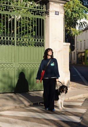 Lässiger navyfarbener Pullover mit grünem Logo, schwarze gestreifte Hose und blaue Schuhe; einen Hund an der Leine; grünes, kunstvoll verziertes Tor im Hintergrund.