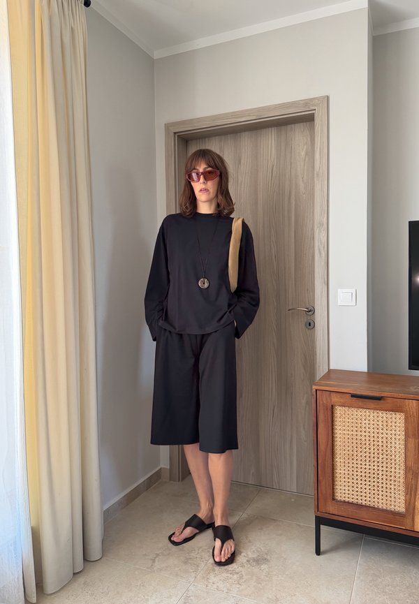Femme portant un haut noir à manches longues et un short avec des lunettes de soleil et des sandales, debout à l'intérieur près d'une porte et d'une armoire en bois.
