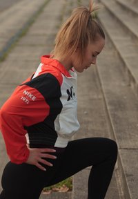 Mlada ženska v rdeči, beli in črni Nike jopici razteguje eno nogo na zunanjih betonskih stopnicah med vadbo.