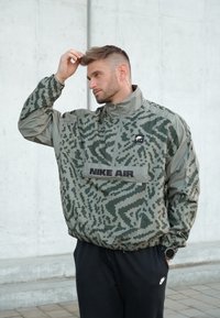 Veste camouflage verte avec une fermeture éclair partielle, des poignets élastiques et une poche avant portant le texte "NIKE AIR". Le matériau semble léger et texturé.