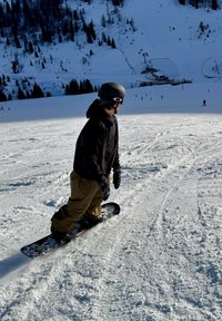 Snowboarder sur la neige portant une veste noire, un pantalon beige et un casque. La planche présente des motifs graphiques, glissant sur une pente enneigée.