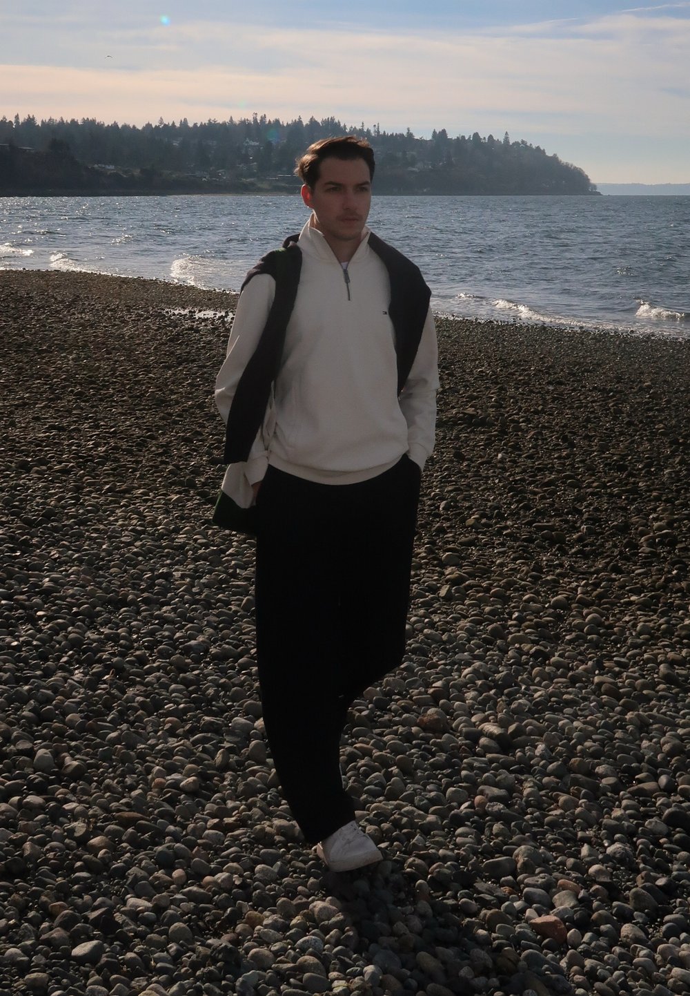 Jeune homme portant un pull blanc et un pantalon noir marchant sur une plage rocheuse près d'une mer calme sous un ciel partiellement nuageux.