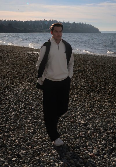 Joven con sudadera blanca y pantalones negros caminando por una playa rocosa cerca del mar tranquilo bajo un cielo parcialmente nublado.
