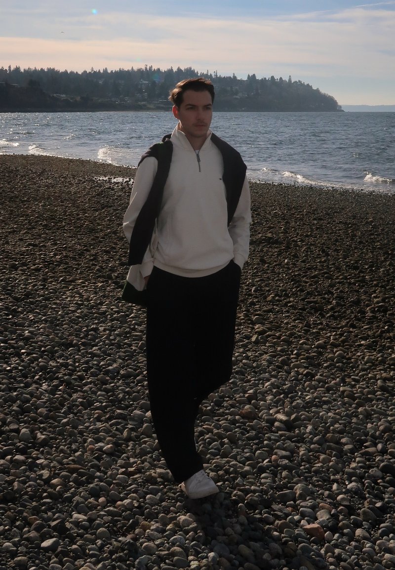 Joven con sudadera blanca y pantalones negros caminando por una playa rocosa cerca del mar tranquilo bajo un cielo parcialmente nublado.