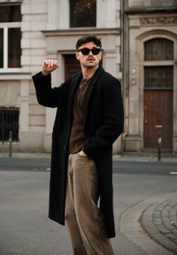 Manteau en laine noire, pull marron et pantalon en velours côtelé beige. Accessoires comprenant des lunettes de soleil noires et des bijoux en or. Debout en plein air.