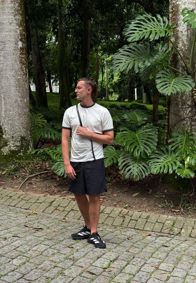 Hombre con camisa blanca, pantalones cortos negros y zapatillas de deporte de pie en un camino de piedra rodeado de grandes hojas verdes y árboles altos en un bosque.