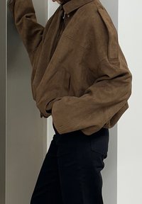 Veste surdimensionnée en suédine marron avec col montant, fermeture éclair et manches larges, associée à un pantalon noir ajusté.