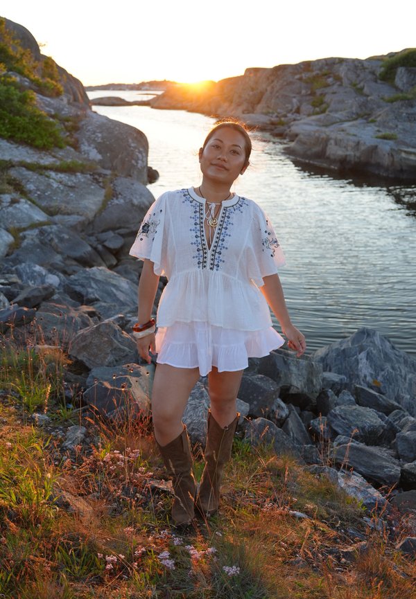 Femme portant une robe blanche et des bottes marron debout sur un rivage rocheux avec de l'eau et un coucher de soleil en arrière-plan.