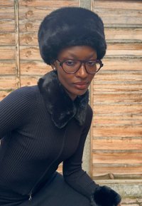 Čierna klobúk z umelej kožušiny, čierny rebrovaný odev s golierom a manžetami z umelej kožušiny, veľké obdĺžnikové okuliare, hladká pokožka, drevené pozadie.