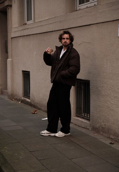 Chaqueta de plumón marrón con cremallera, camiseta blanca, pantalones negros y zapatillas blancas. Sosteniendo una taza beige, de pie contra una pared texturizada.