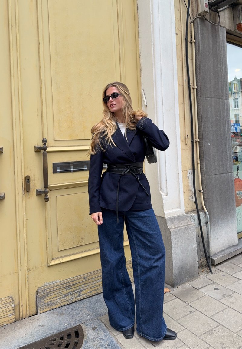 Blazer azul-marinho com lapelas largas e um cinto preto, combinado com jeans azuis oversized. Óculos de sol pretos e uma pequena bolsa completam o visual.