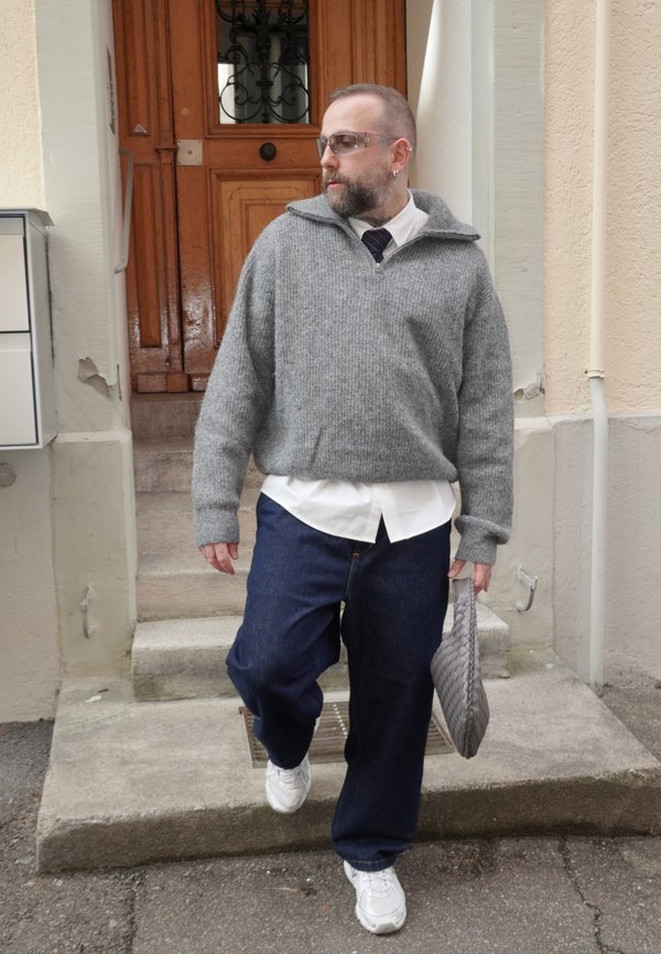 Mann iført grå genser, hvit skjorte, blå jeans, hvite joggesko og solbriller går ned betongtrapper mens han holder en grå vevd clutchveske.