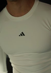 Valge pika varrukaga spordisärk, millel on lõhestatud disain. Rinnal on must Adidas logo. Sile ja veniv materjal.