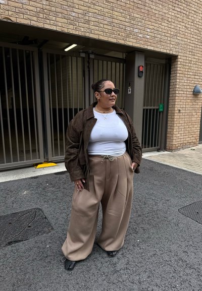 Chaqueta de ante marrón sobre una camisa blanca, combinada con pantalones anchos beige y zapatos negros. Los accesorios incluyen gafas de sol y un collar de perlas.