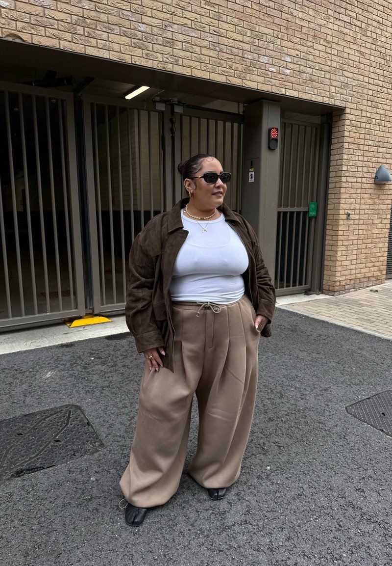 Veste en daim marron portée sur une chemise blanche, associée à un pantalon beige à jambes larges et des chaussures noires. Accessoires : lunettes de soleil et collier de perles.