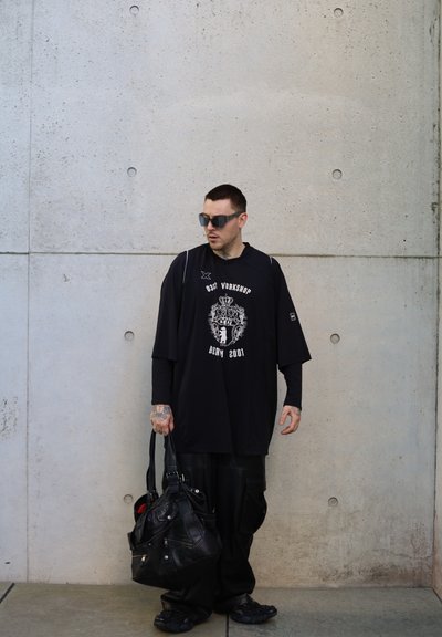 Camisa negra de gran tamaño con gráfico y texto en blanco, combinada con pantalones negros holgados. Bolso de cuero negro y gafas de sol rectangulares, sobre un fondo de concreto.
