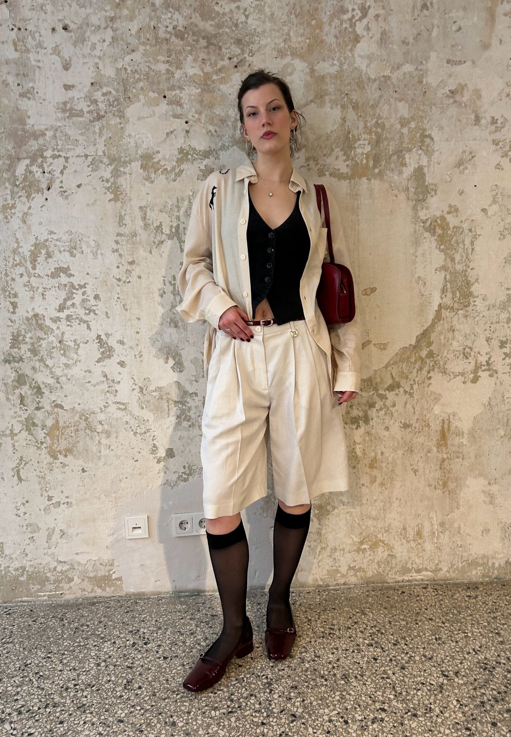 Kreminės spalvos per didelė marškinė, juodas cropped topas, klostuoti šortai, juodos pėdkelnės ir bordo spalvos batai. Raudona rankinė ir minimalūs auskarai.