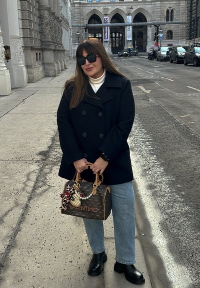 Abrigo negro tipo peacoat, suéter blanco de cuello alto, jeans de mezclilla azul claro y botas negras de tobillo. Bolso Valentino marrón con charm decorativos. Entorno urbano.