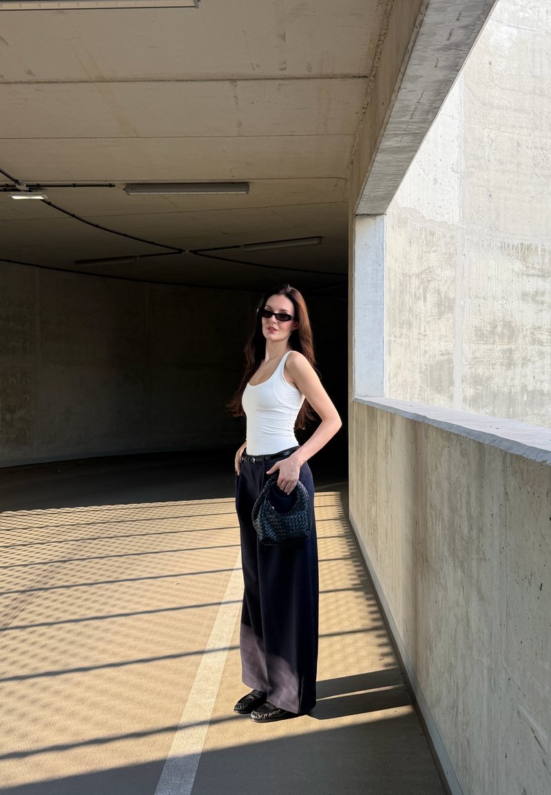 Donna che indossa un top bianco, pantaloni neri larghi, occhiali da sole, tiene una borsa nera, in piedi in un parcheggio in cemento illuminato dal sole con ombre sul pavimento.