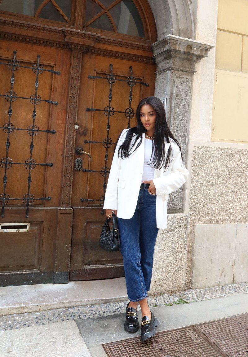 Λευκό oversized σακάκι, εφαρμοστό λευκό τοπ, μπλε cropped τζιν, μαύρες πλατφόρμες με χρυσές λεπτομέρειες, κρατώντας μια υφασμάτινη μαύρη τσάντα. Φόντο ξύλινη πόρτα.