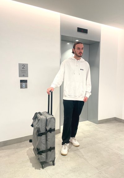 Maleta gris con ruedas y correas negras, de forma cuadrada, material de tela, apoyada sobre un suelo de baldosas cerca de un ascensor.