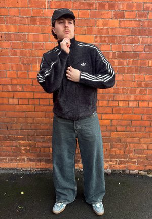 Chaqueta de chándal negra y peluda con rayas blancas, combinada con jeans oscuros y sueltos y zapatillas claras, sobre un fondo de pared de ladrillos rojos.