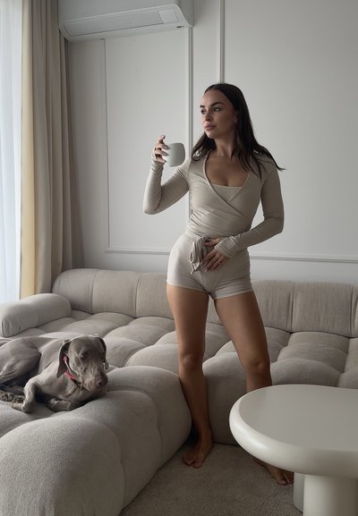 Mujer con ropa cómoda beige sosteniendo una taza blanca, de pie descalza junto a un sofá gris claro con un perro tumbado sobre él en un salón minimalista.