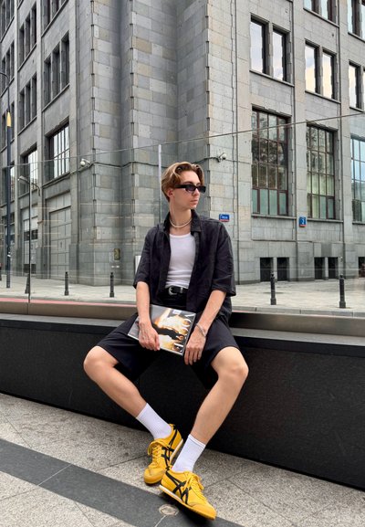 Camisa negra de manga corta sobre una camiseta blanca, pantalones cortos negros, zapatillas amarillas, sosteniendo una revista, sentado sobre una superficie oscura. Fondo urbano.