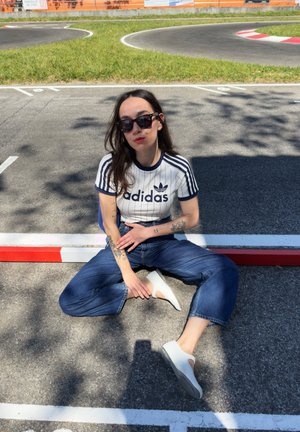 Balta Adidas marškinėliai su tamsiai mėlynomis juostelėmis ir logotipu, aukštu liemeniu mėlynos džinsai, balti batai, juodi saulės akiniai, sėdintis ant asfalto su raudonais ir baltais žymėjimais.
