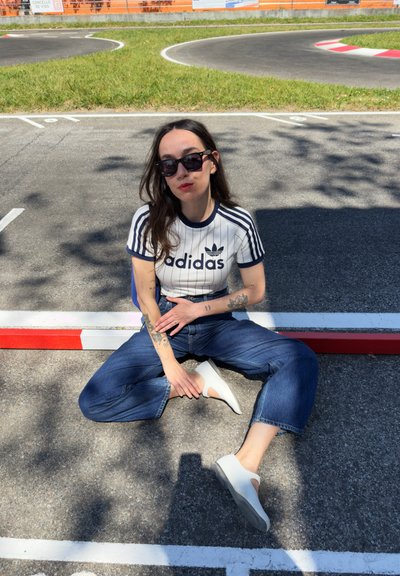 Camiseta blanca de Adidas con rayas y logotipo en navy, jeans azul de cintura alta, zapatos blancos, gafas de sol negras, sentado sobre asfalto con marcas rojas y blancas.