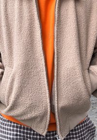 Μπεζ φλις ζακέτα με φερμουάρ και πλαϊνές τσέπες πάνω από πορτοκαλί πουκάμισο; διαθέτει ριμπ μανσέτες και στρίφωμα, συμπληρωμένα από καρό παντελόνι.