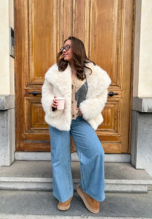 Mujer con gafas, vestida con un abrigo de piel blanco, suéter beige, vaqueros anchos azules y zapatos marrones, sosteniendo una taza y posando frente a una puerta de madera.