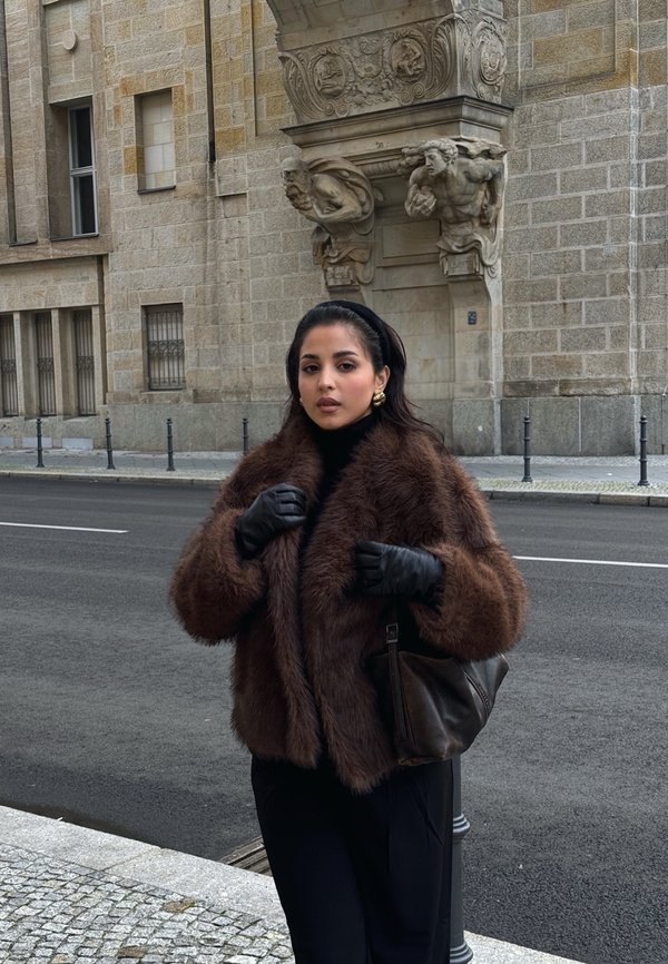 Mujer con abrigo de piel marrón y guantes negros está en la calle de la ciudad cerca de un edificio de piedra con esculturas detalladas en el arco superior.