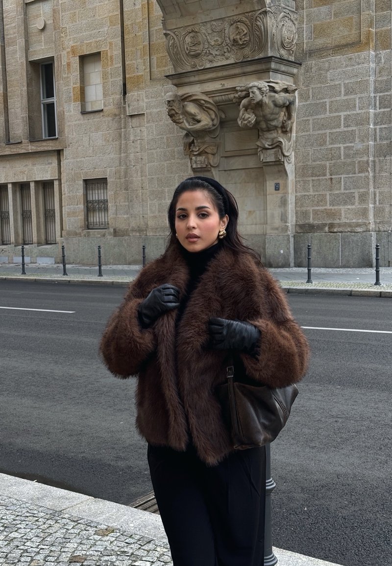 Femme en manteau de fourrure marron et gants noirs se tient dans une rue de la ville près d'un bâtiment en pierre avec des sculptures détaillées sur l'arc au-dessus.