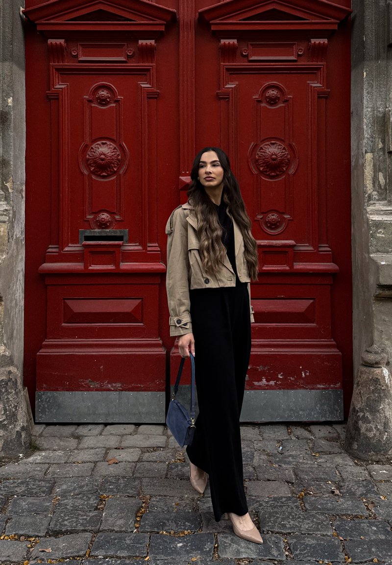 Femme aux longs cheveux foncés, vêtue d'une veste beige et d'une tenue noire, tenant un sac à main bleu, debout dans une rue pavée près de grandes portes décoratives rouges.
