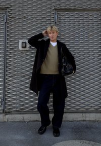 Una persona con indosso un cappotto nero sopra un maglione verde, jeans scuri e scarpe nere, che tiene in mano una borsa nera, è in piedi davanti a uno sfondo grigio testurizzato.